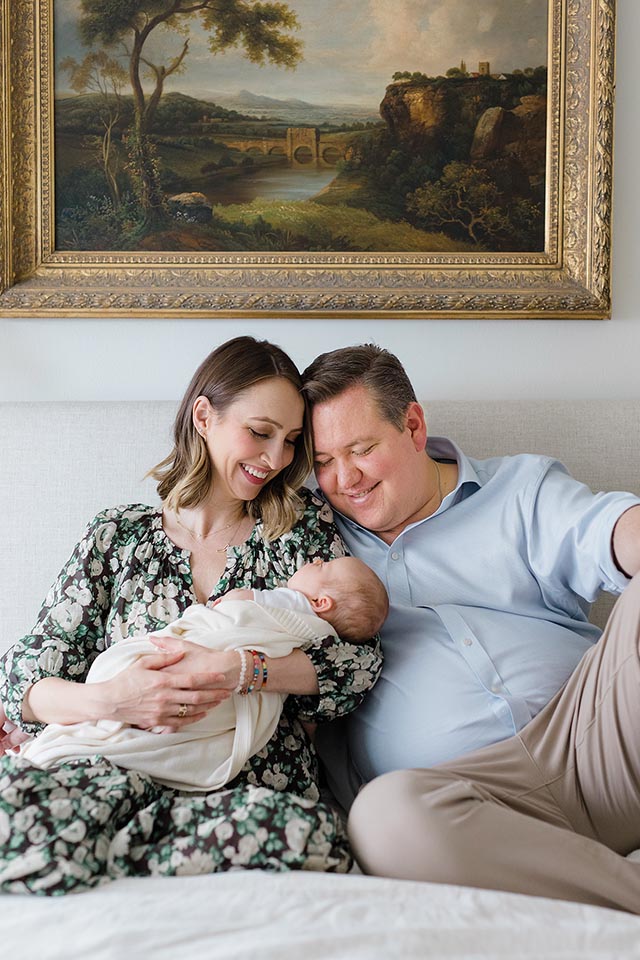 Colgate couple sits on bed under portrait, looks down lovingly at their newborn