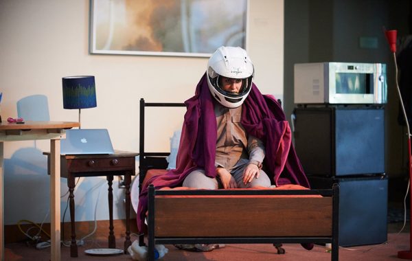 Dani Solomon ’13 performing One Way Red, sitting on the end of a bed wearing a motorcycle helmet and draped in a blanket.