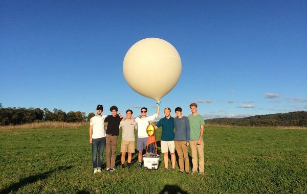 Seven students pose in a field with a weather balloon attached to a camera-housing apparatus