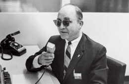 Michael Supa wearing sunglasses and talking into a table microphone