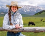 Faith Hamlin standards against a post fence with cows grazing and mountains in the background