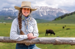 Faith Hamlin leans against post on her ranch with open land, animals and mountains in the background.