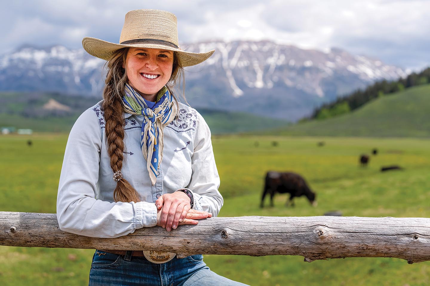 Faith Hamlin leans against post on her ranch with open land, animals and mountains in the background.