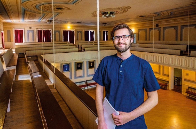 Eli Watson ' 24 hold papers while standing on second floor of Oneida Community Mansion House. The picture shows details of the house and a view to the main level below.