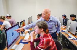 Professor Jeff Bary points to a computer monitor while a student looks on. Other students are working on computers in the background.