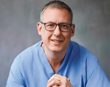 Headshot of Marc David Munk, wearing blue hospital scrubs