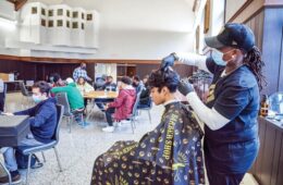 Male student of color wearing a barber shop cape, having his hair cut by a woman of color. In the background, male students of color are sitting at tables in groups interacting.