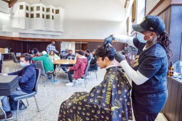Male student of color wearing a barber shop cape, having his hair cut by a woman of color. In the background, male students of color are sitting at tables in groups interacting.