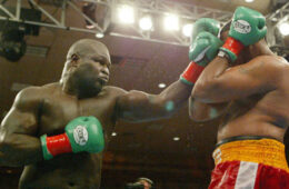 James ”Lights Out“ Toney, wearing green boxing gloves, punching an opponent holding his forearms and gloves up to cover his neck and face