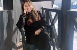 Portrait of Linda Wang, seated on a rustic porch with a high, black-painted railing, smiling, with long, straight hair hanging down, her chin resting on one elbow,with snow-covered mountains in the background