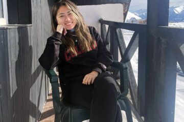 Portrait of Linda Wang, seated on a rustic porch with a high, black-painted railing, smiling, with long, straight hair hanging down, her chin resting on one elbow,with snow-covered mountains in the background