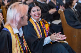 Alex Tran and Hilary Almanza, both wearing graduation robes, gold stoles, and multiple cords, smile at each other while seated in Memorial Chapel at the Baccalaureate Service