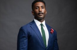 Portrait of Wesley Morris wearing a navy suit with red floral boutinier, white shirt, and green tie.gray background
