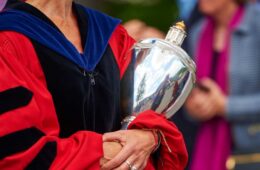 Woman, seen from the chin down in red, black, and blue academic regalia, holding Colgate University's mace