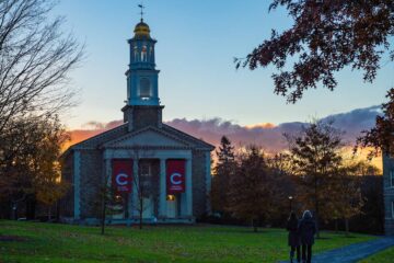 Colgate Memorial Chapel at sunset with low bank of pink clouds behind it. Two people walking in the foreground.