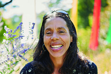 Woman of color with long straight hair and glasses on top of her head smiles in an outdoor setting. Delicate purple flowers to her left.