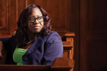 African-American woman with long, wavy, dark red hair wearing a lime green top and navy blazer and navy glasses, sits in the dark wooden pews of a church, looking off to the right