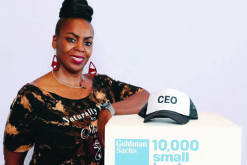 Shirene Brown, an African-American woman leans with her elbow on a white box labelled Goldman Sachs: 10,000 small busineses. The box has a black and white baseball cap that says CEO. She wears red woodcut dangling earrings and her hair is piled in a bun on top of her head.