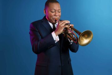 Wynton Marsalis, playing his trumpet in front of a blue studio backdrop