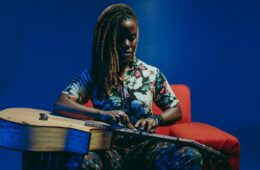 Guitarist Yasmin Williams, in long dreadlocks to one side and a flowered top, taps the strings on the guitar on her lap, sits on a rust colored easy chair with a dark blue backdrop.