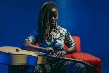 Guitarist Yasmin Williams, in long dreadlocks to one side and a flowered top, taps the strings on the guitar on her lap, sits on a rust colored easy chair with a dark blue backdrop.