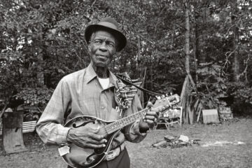 Older black man in a hat and playing a mandolin. Black and white image.