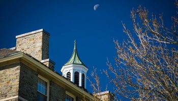 A daytime moon rises over McGregory Hall.