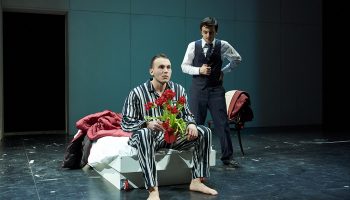 A student actor in a suit on stage speaks with another actor in pajamas holding a vase of red flowers.