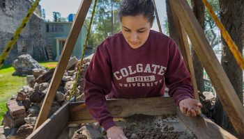 Female student in a Colgate sweatshirt uses a sifter in the field during research