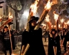 Men and boys process at night, dressed in black, holding two large bundles of torches, one on each shoulder